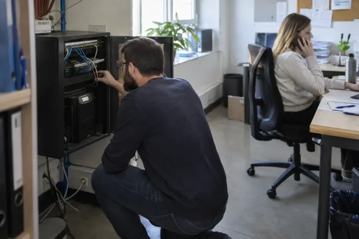 Technician checking network backup equipment while an office employee uses a business phone