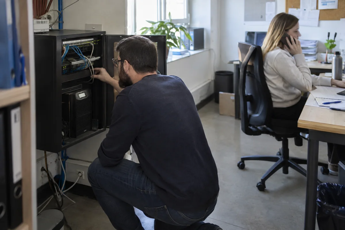 Technician checking network backup equipment while an office employee uses a business phone